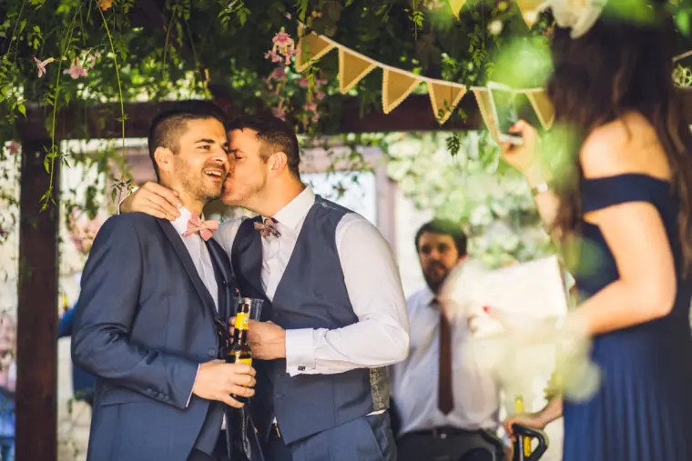 Groom in a blue suit kissing his best man on the cheek under the bunting