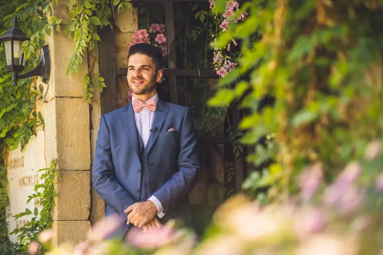 Groom in a blue suit and pink bow tie standing in the ivy-covered archway