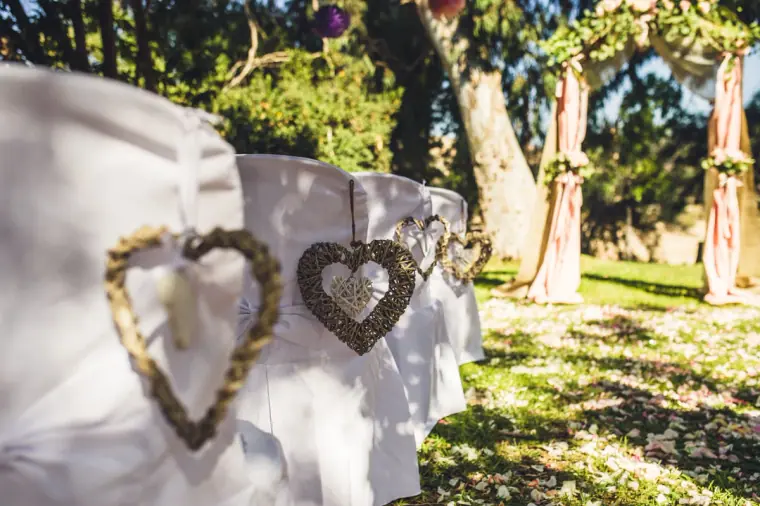 Woven heart decorations on white-draped ceremony chairs in the sunny garden