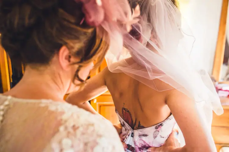 Guest in a fascinator helping the bride into her floral gown