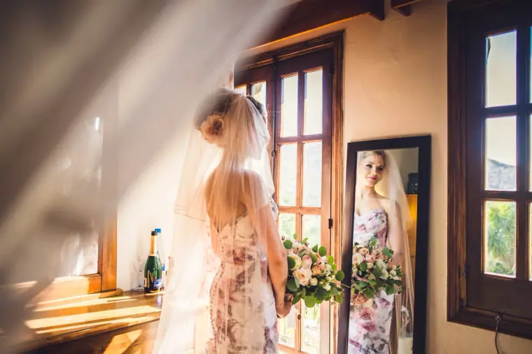 Bride in a floral gown and veil seen from behind, reflected in a bedroom mirror