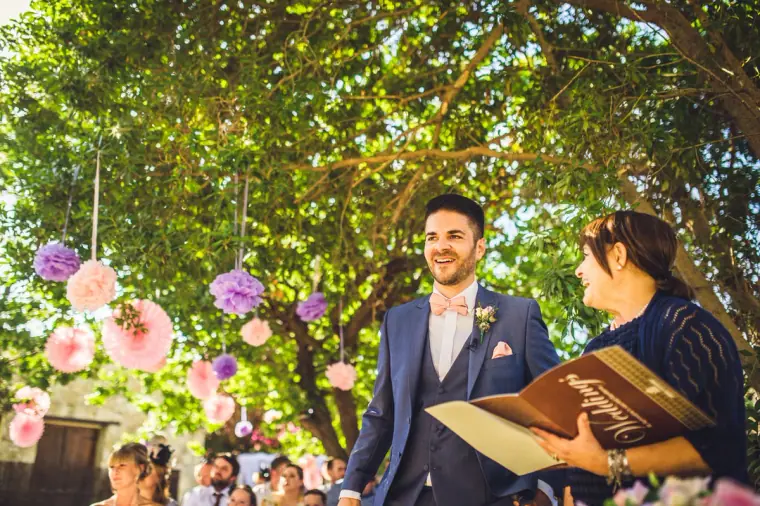 Smiling groom beside the celebrant holding a wooden-covered ceremony book under paper pompoms