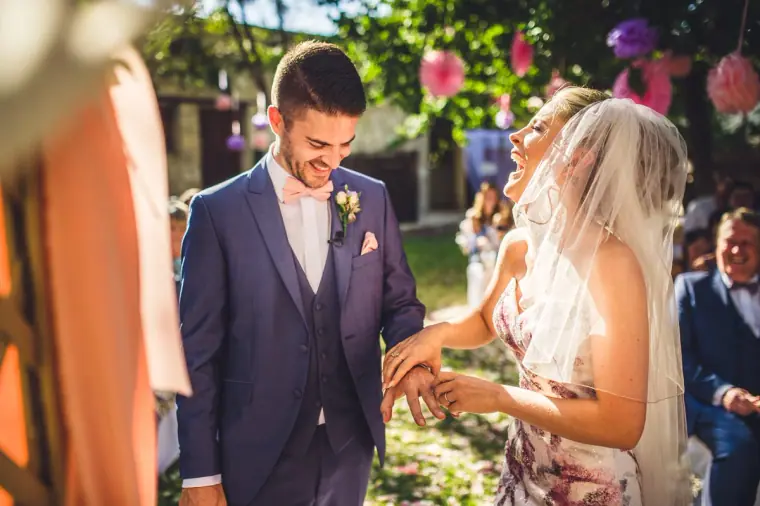 Groom smiling as he slides on the bride's ring beneath pink pompoms