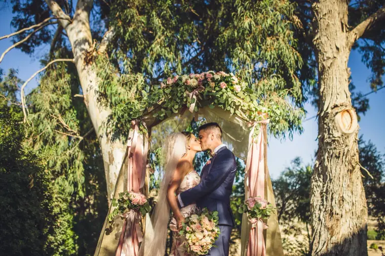 Bride and groom kissing beneath tall trees by the draped ceremony arch
