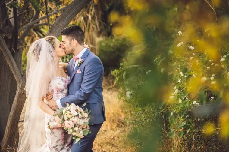 Groom kissing the bride's forehead as she holds her bouquet among the trees