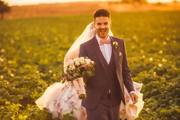 Groom laughing as he walks through the field with the bride behind him