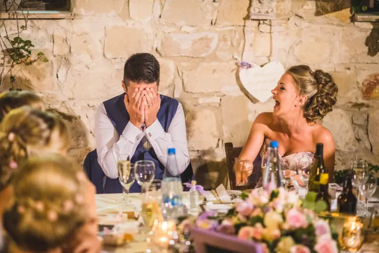 Groom covering his face in embarrassment as the bride laughs during the speeches