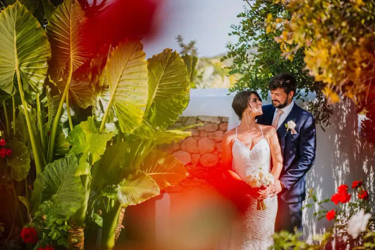 Bride and groom gazing at each other framed by tropical leaves and red flowers