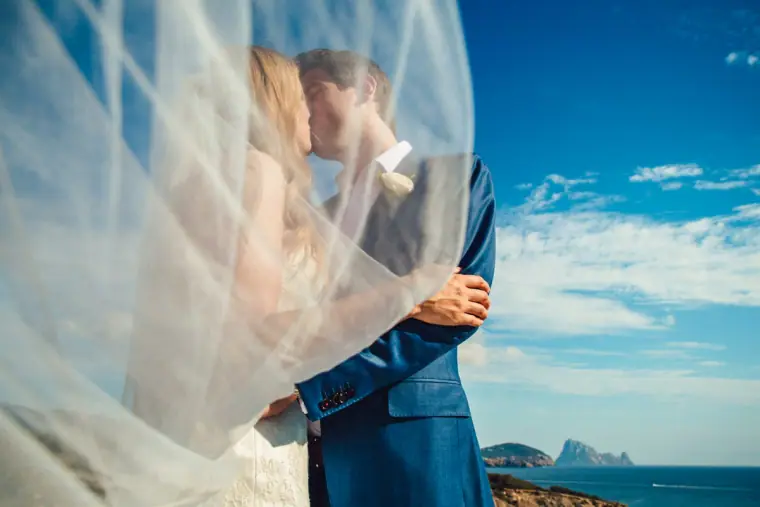 Couple kissing wrapped in a billowing veil with the sea and a rocky island off Ibiza behind