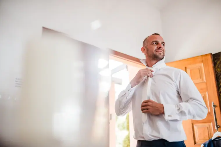 Groom buttoning his white shirt by a sunny window