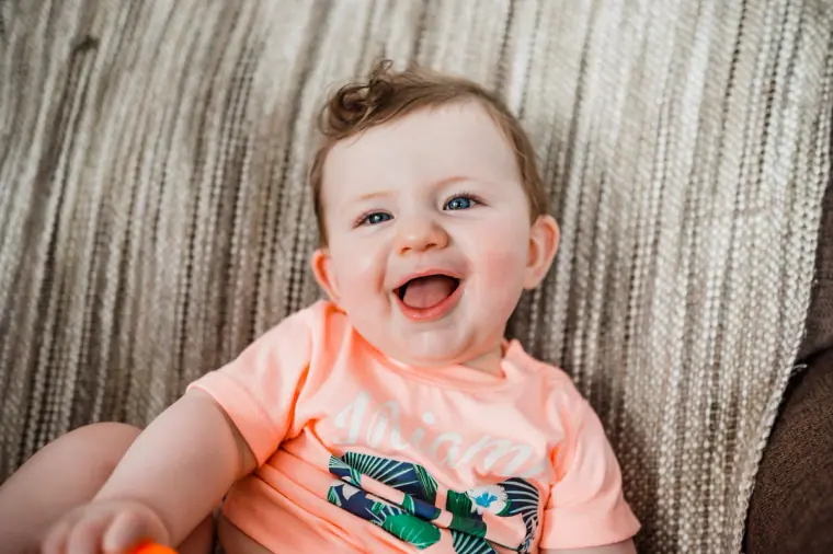 Laughing baby in a coral T-shirt sitting on a sofa