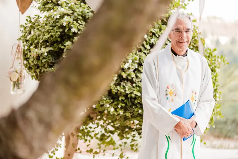 Priest in white vestments holding a blue book beneath the trees