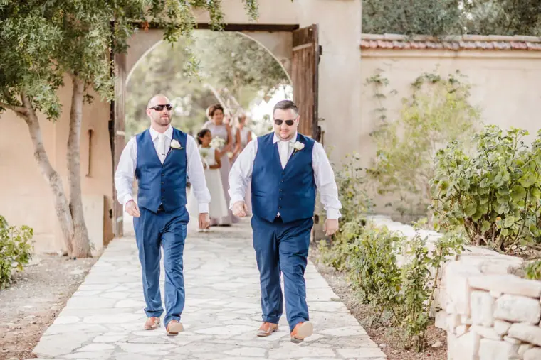 Two groomsmen in navy waistcoats walking down the stone path towards the ceremony