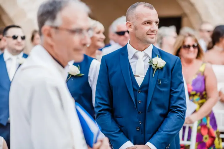 Groom in a navy three-piece suit smiling beside the priest as he waits for the bride
