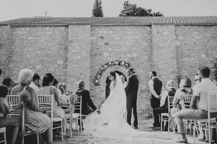 Black-and-white photo of the couple before the floral-framed monastery door, guests seated either side