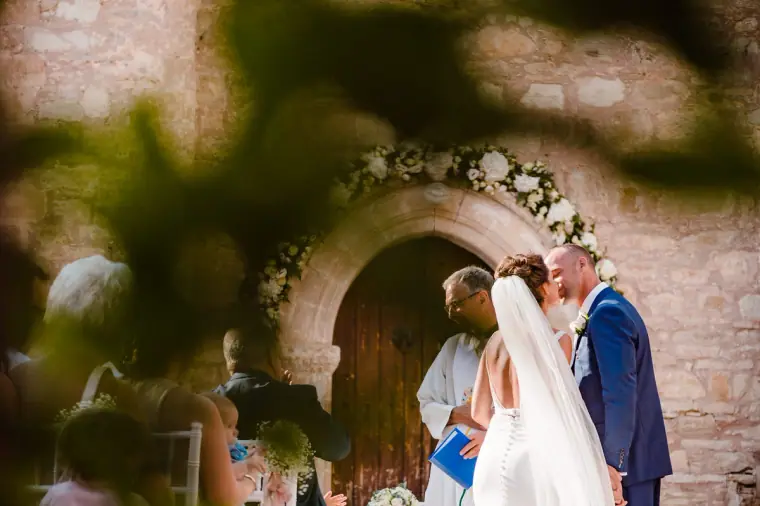 Couple's first kiss before the arched chapel door, framed by soft greenery