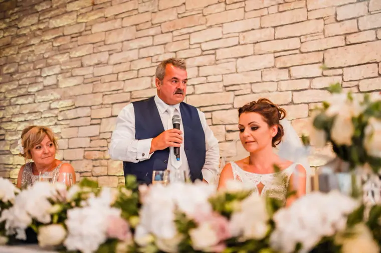 Man in a navy waistcoat giving a speech as the bride smiles beside him