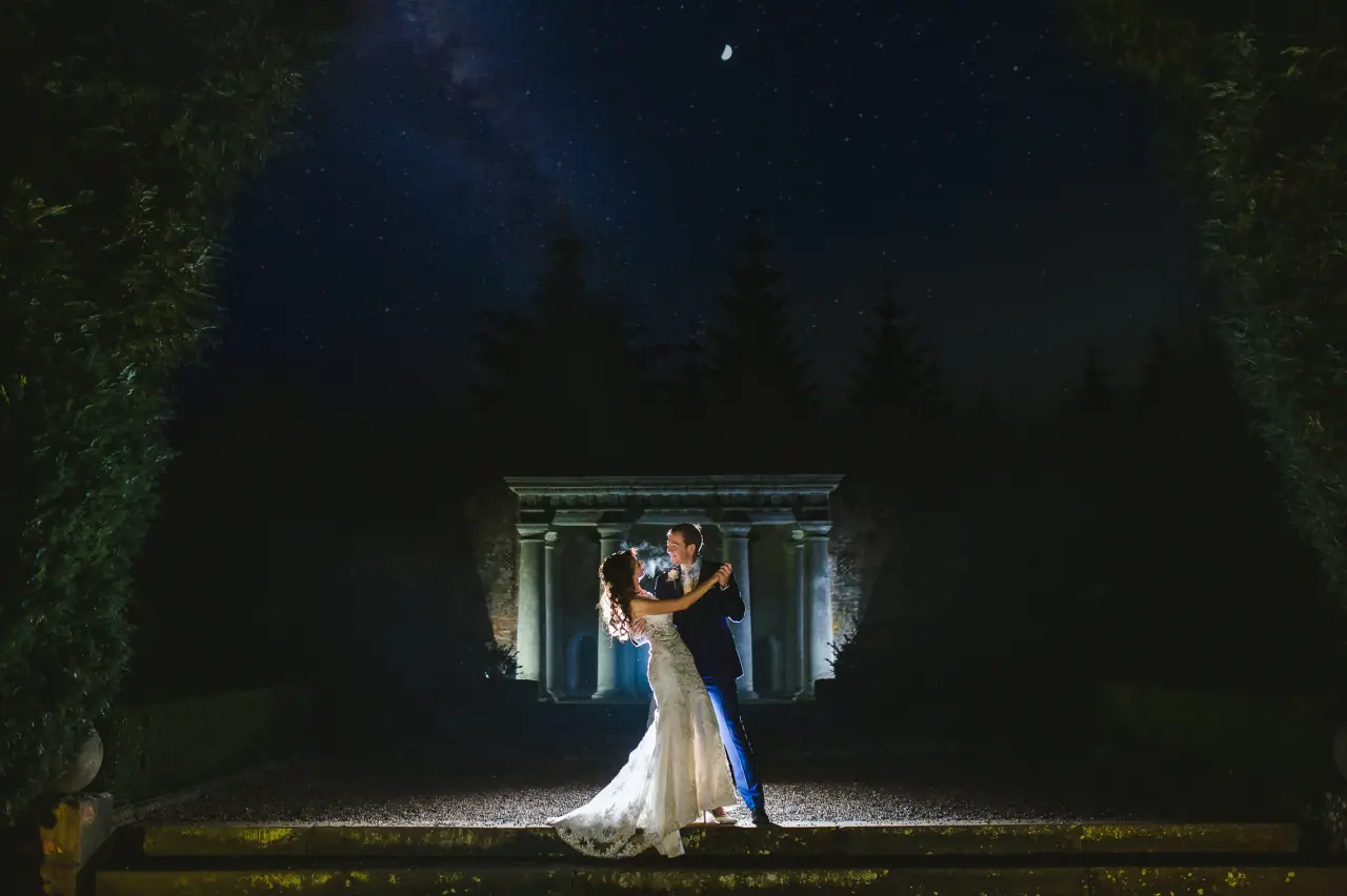 Elina and Iain embracing before a lit stone temple at night at Wotton House, Dorking
