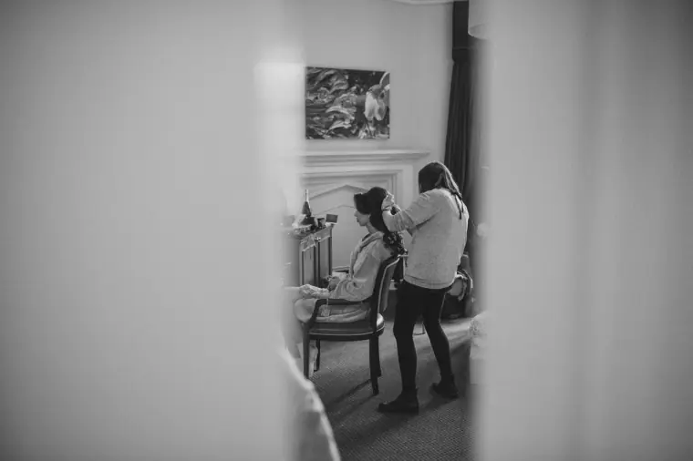 Black-and-white photo of the bride having her hair styled, seen through a doorway