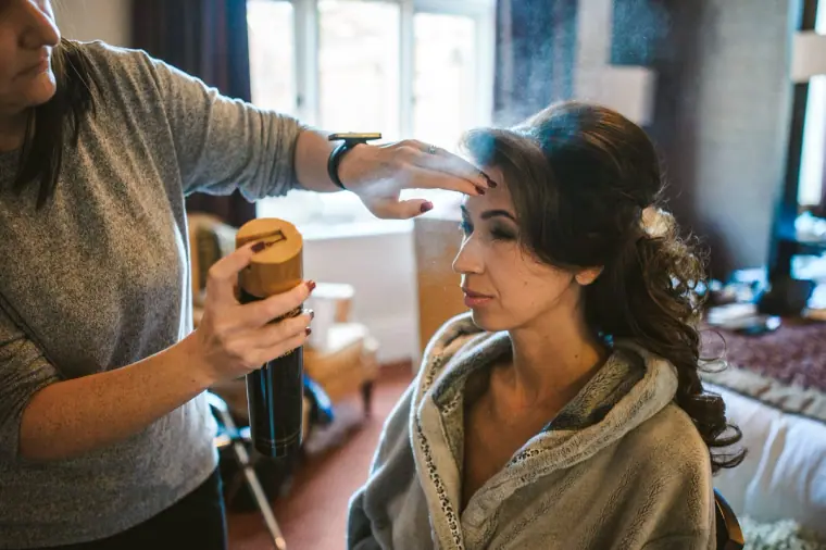 Hairstylist setting the bride's hair with spray as she sits in a grey robe