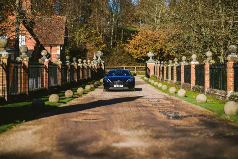 Dark car driving up the tree-lined drive between brick pillars at Wotton House