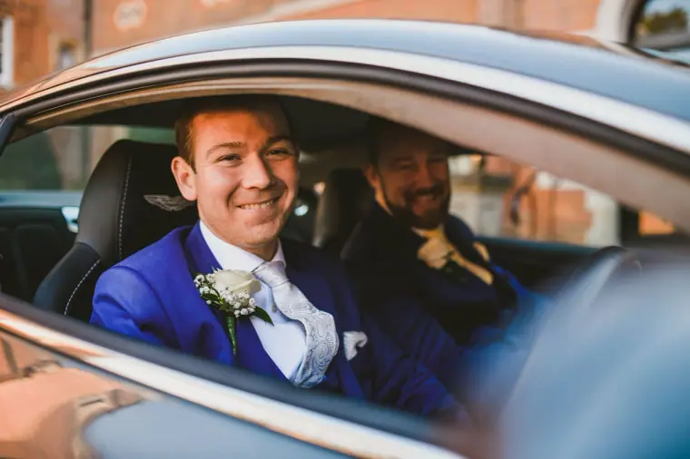 Groom in a blue suit smiling from the passenger seat of the wedding car