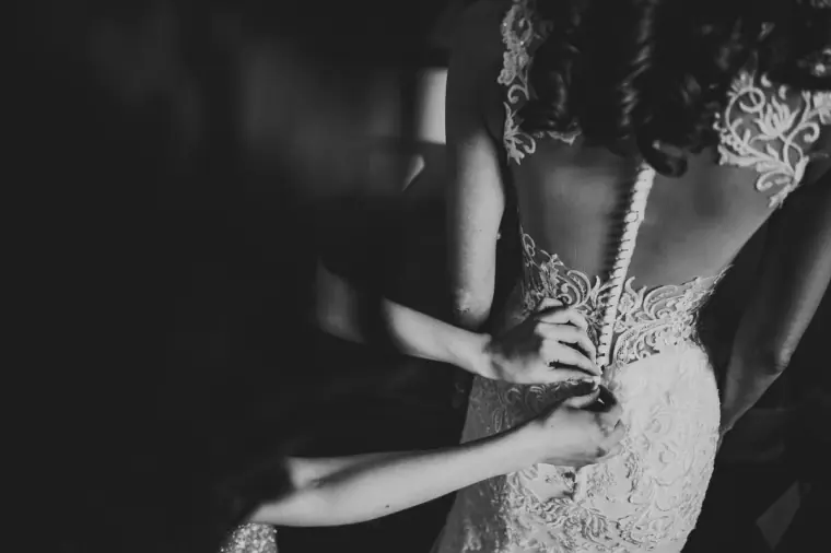 Black-and-white photo of the bride's lace gown being buttoned at the back