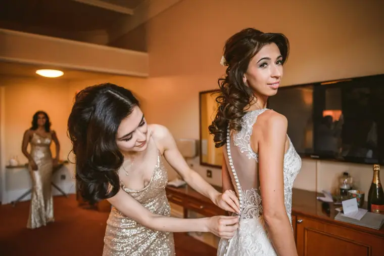 Bridesmaid in gold sequins fastening the back of the bride's lace dress