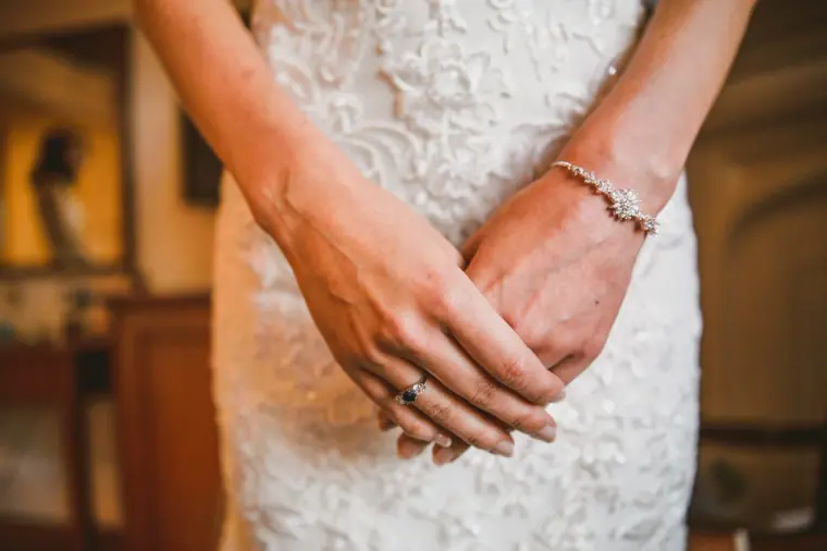 Close-up of the bride's clasped hands, bracelet and ring against her lace gown