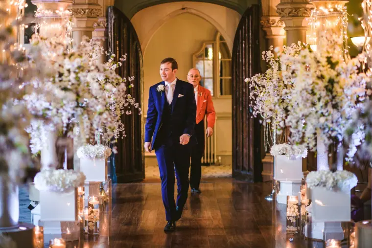 Groom walking down a candlelit aisle lined with white blossom displays at Wotton House