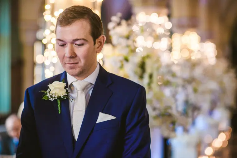 Iain waiting for his bride in a navy suit among white blossom arrangements