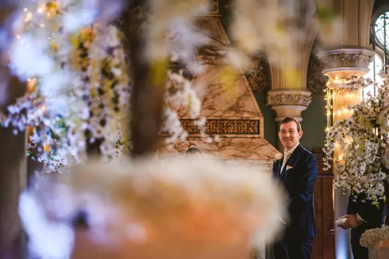 Groom glancing back down the aisle, framed by soft-focus white flowers at Wotton House