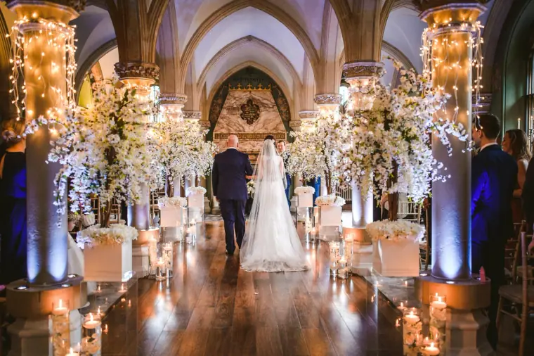 Bride walking with her father down a candlelit aisle under gothic arches at Wotton House