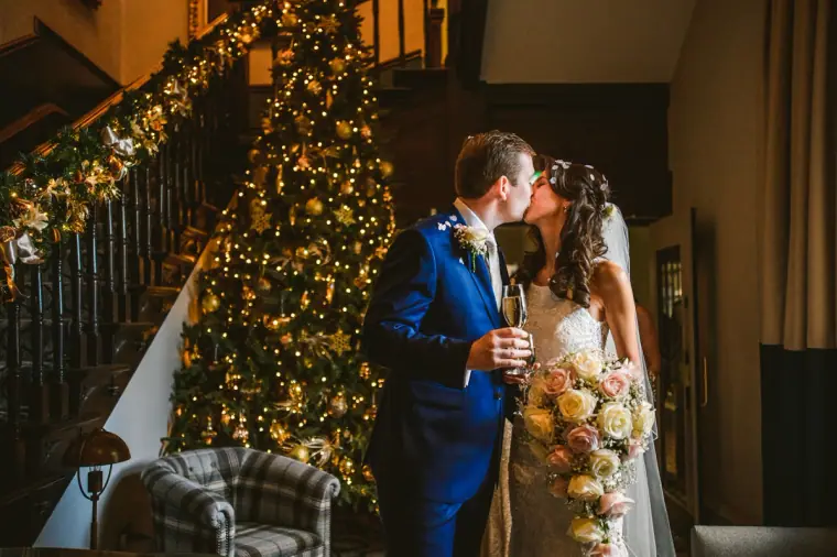 Elina and Iain kissing beside a lit Christmas tree at the foot of the staircase
