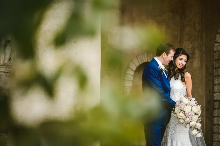 Elina and Iain framed through soft green foliage beside a stone arch