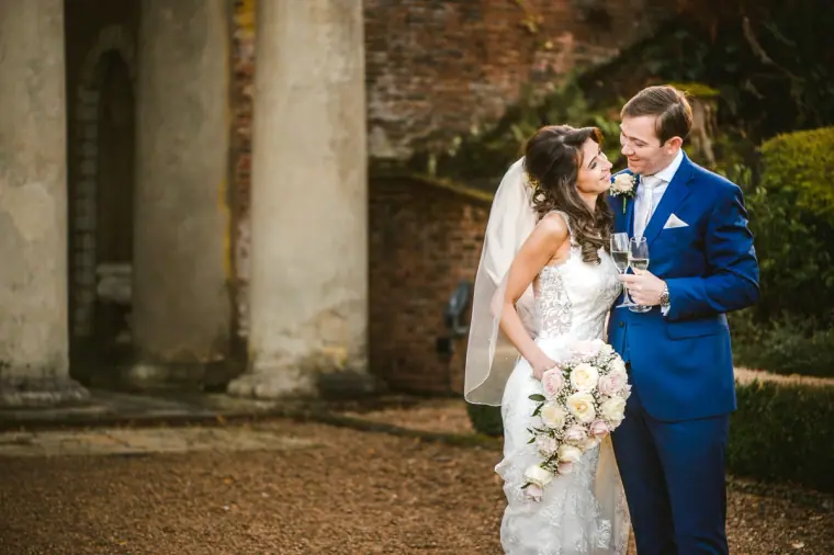 Iain and Elina laughing beside weathered stone columns, her bouquet in hand