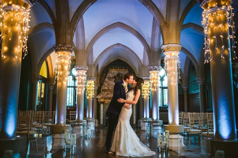 Couple kissing in the empty vaulted ceremony hall lit by fairy-light columns at Wotton House