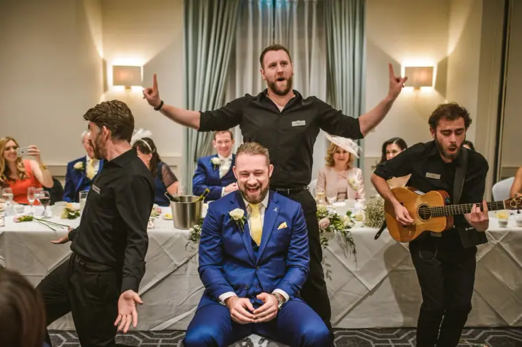 Groomsman laughing as singing waiters perform around him at the reception