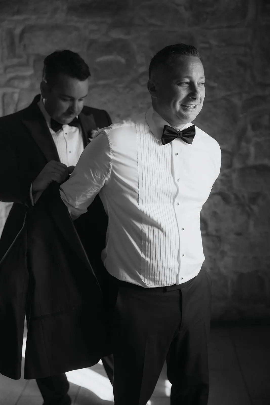 Black-and-white photo of a groomsman helping Oliver into his dinner jacket