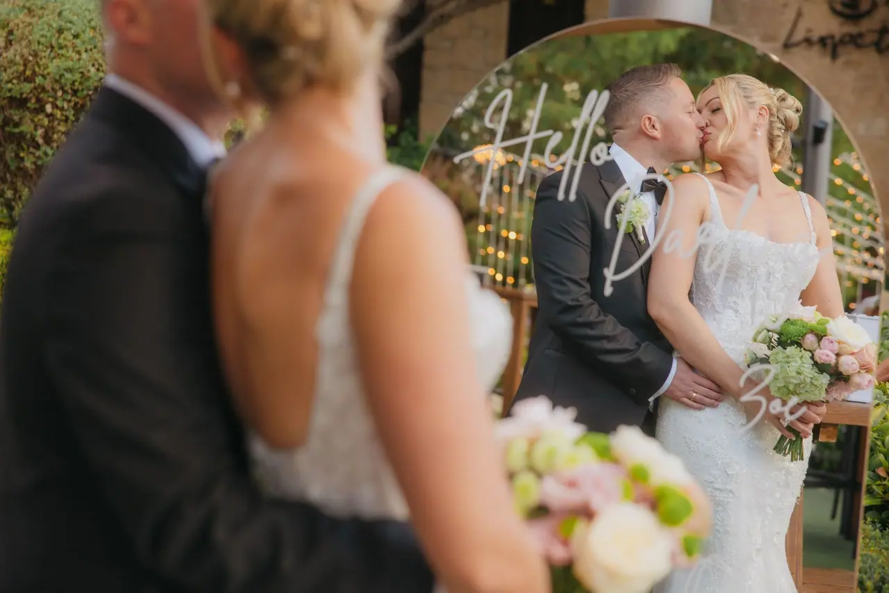 Zoe and Oliver kissing, reflected in a mirrored welcome sign in the Liopetro gardens