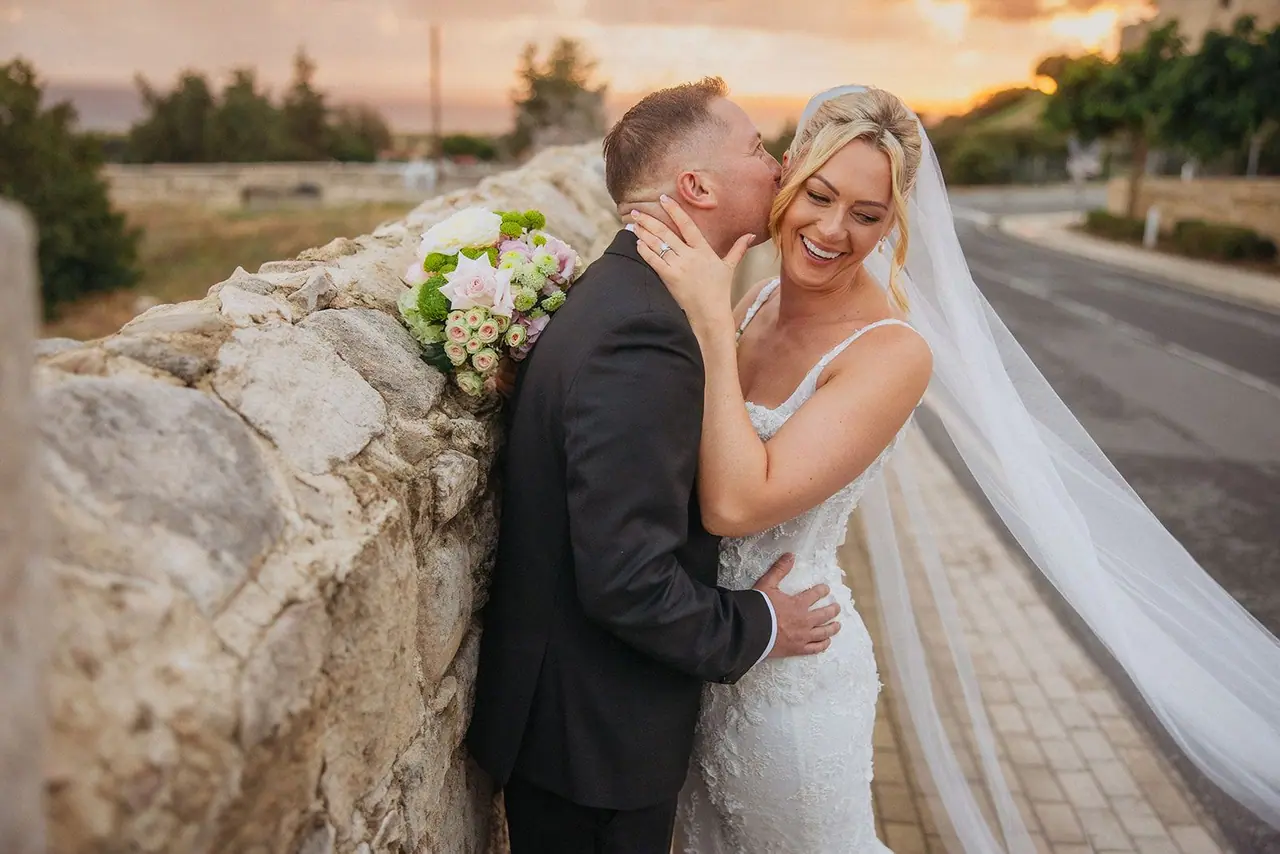 Oliver kissing Zoe's cheek against an old stone wall at sunset near Liopetro