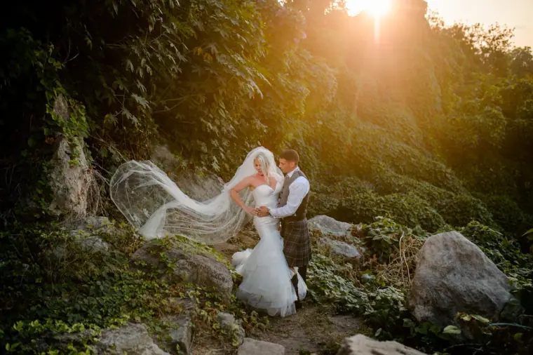 Maryellen's veil billowing as she and Ben embrace among forest rocks in low sun