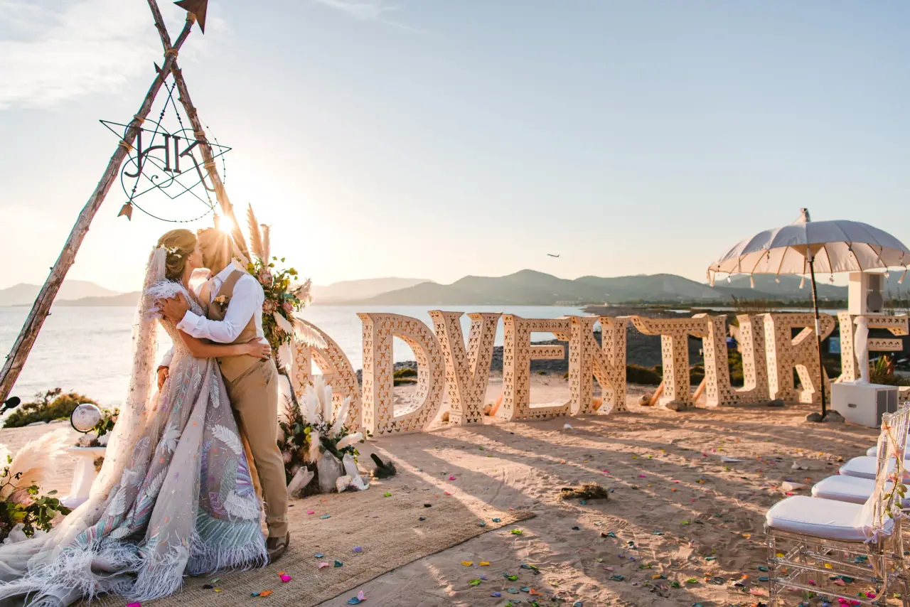 Couple kissing beneath a macramé triangle arch beside giant 'Adventure' letters at sunset by the sea