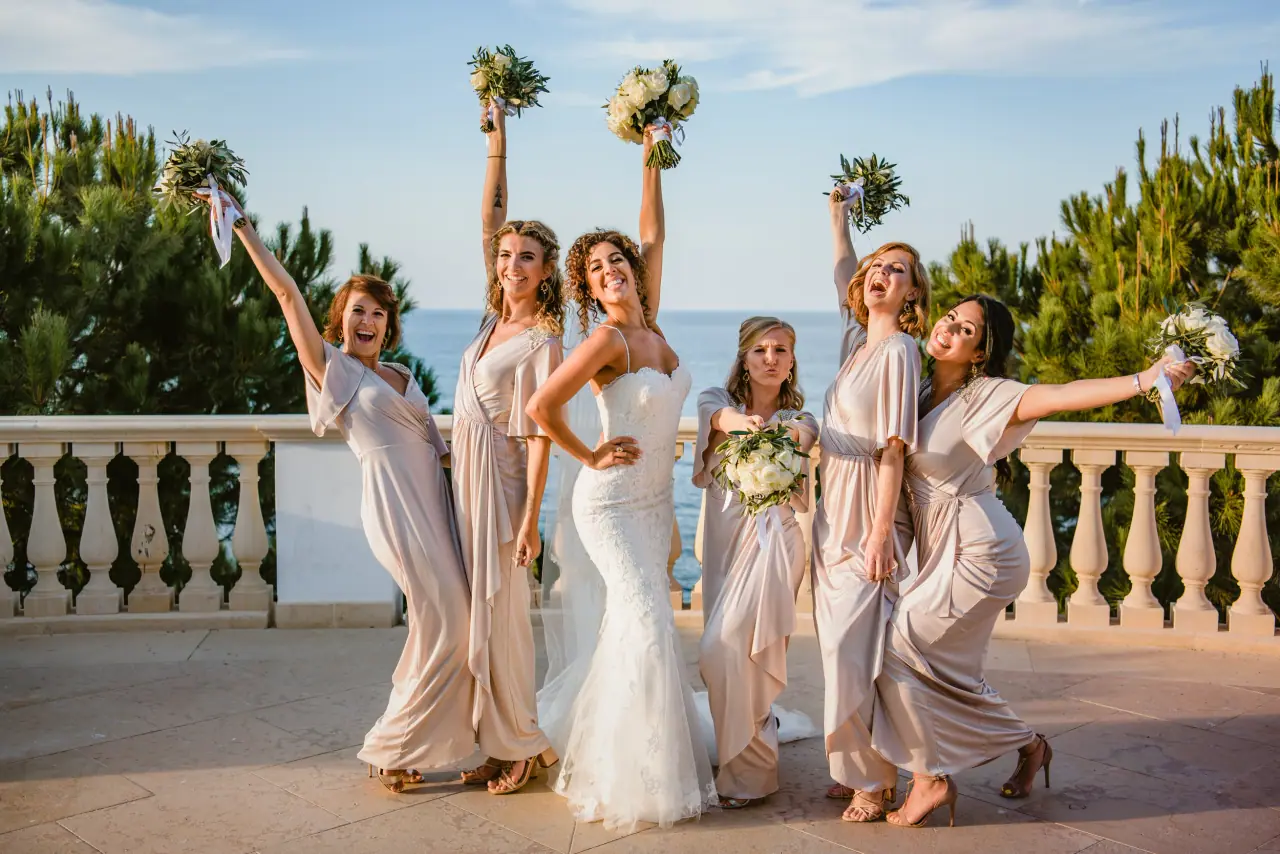 Bride and bridesmaids raising their bouquets on a balustraded terrace above the sea