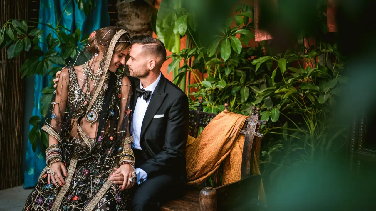 Bride in an embroidered South Asian outfit and groom in a tuxedo seated among tropical plants