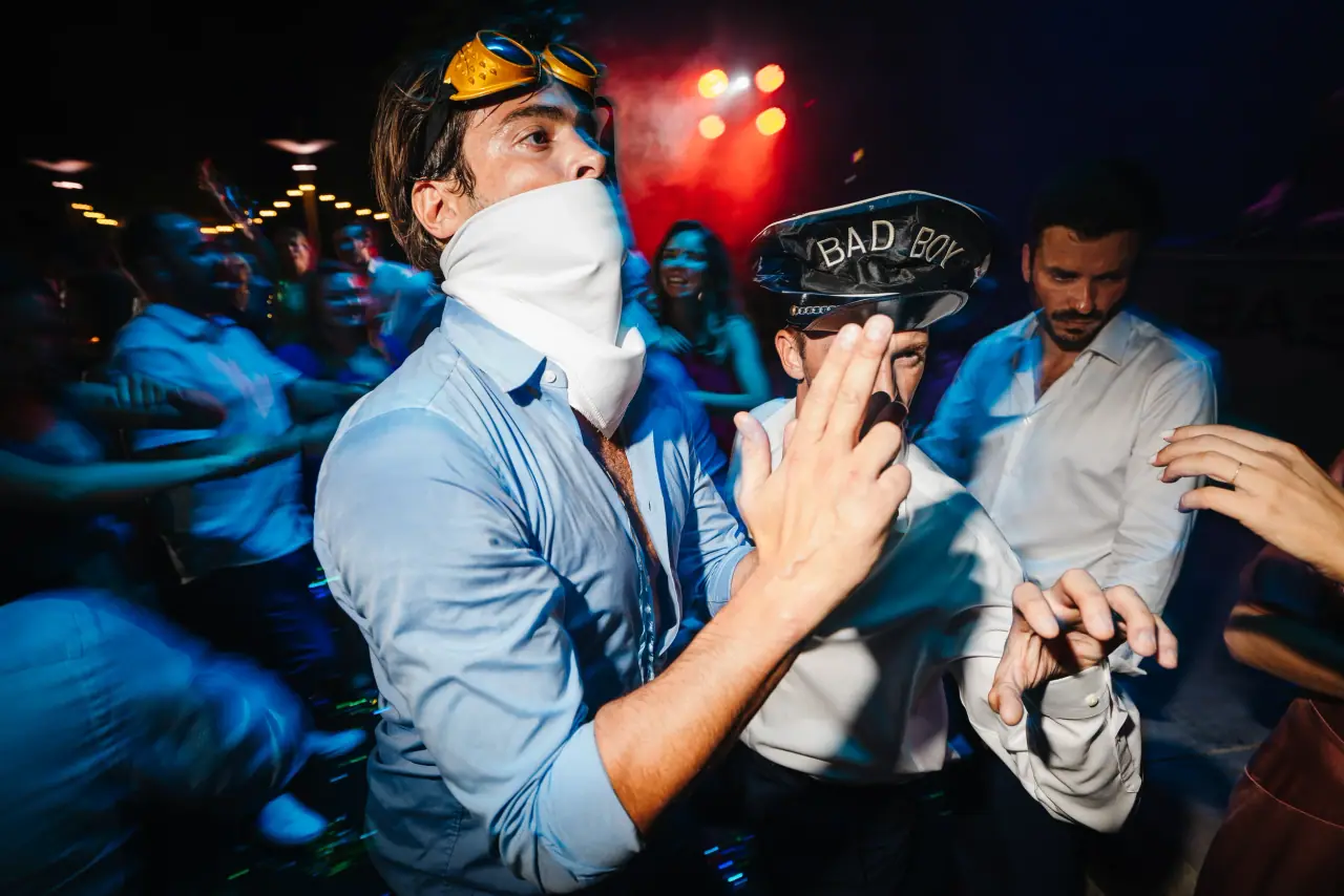 Groomsman with a scarf over his face partying among guests under red lights