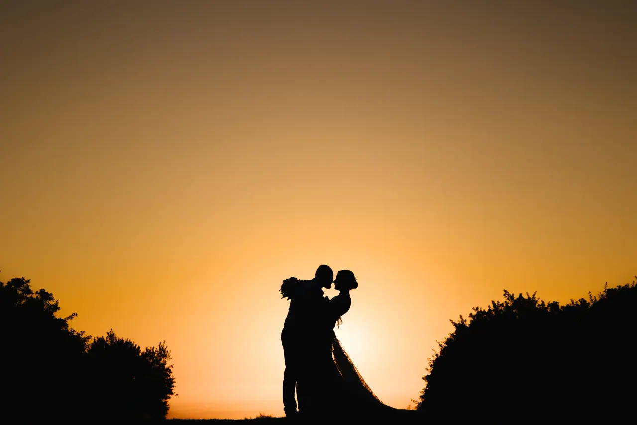 Bride and groom silhouetted in a kiss against an orange sunset sky