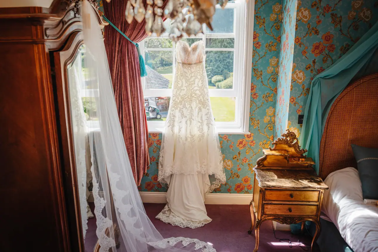 Wedding dress and long veil hanging at a window in a floral-wallpapered bedroom