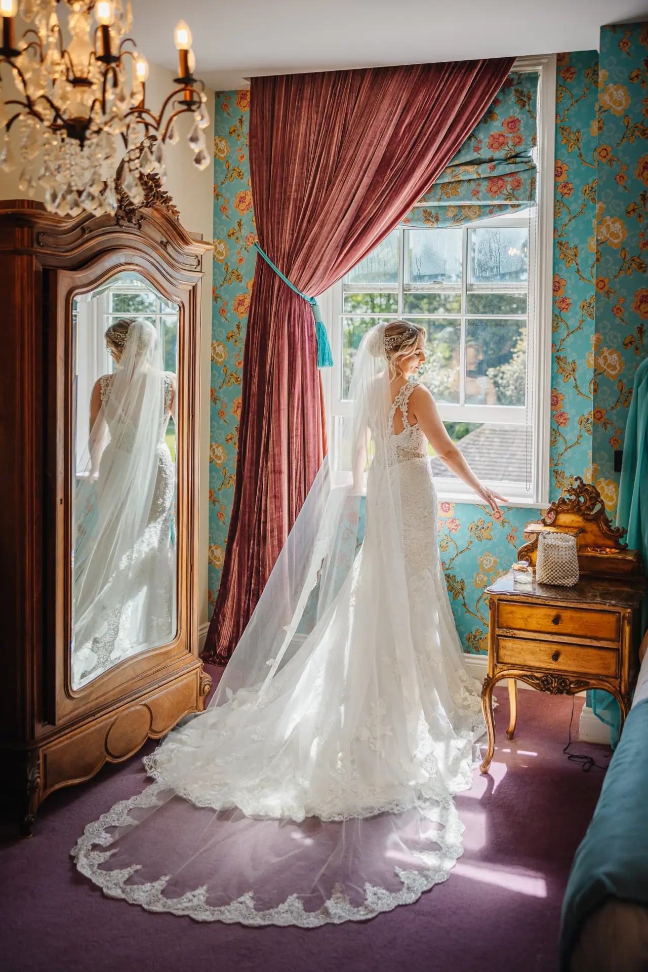 Olivia in her gown and long veil looking out of a curtained window