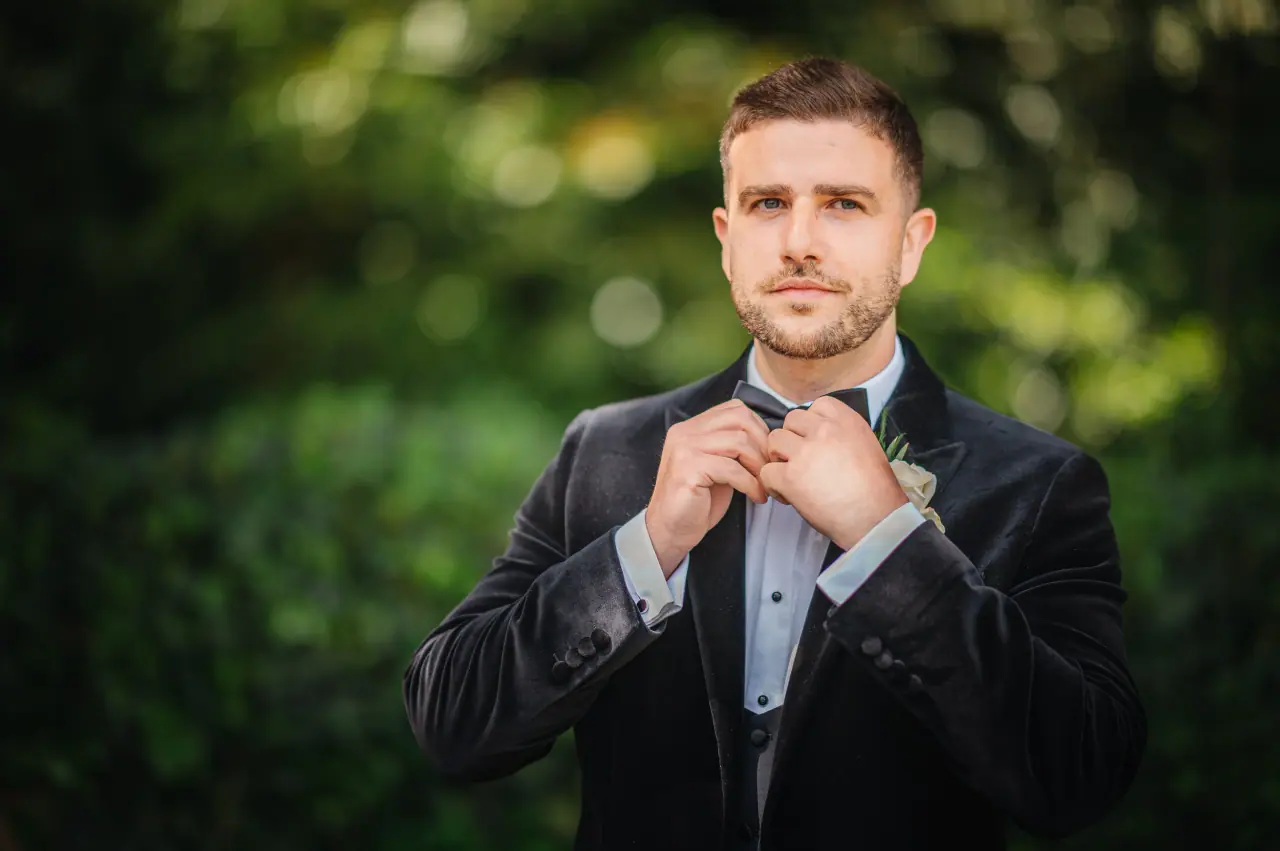 Alex adjusting his bow tie in a black velvet tuxedo against green foliage